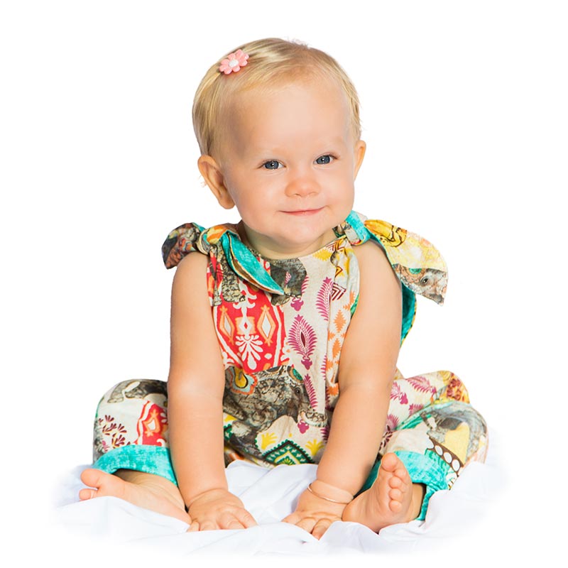 A 12 month blond baby girl sits against a white backdrop as she smiles while wearing an Elephant Walk Romper made by Baby Blue.