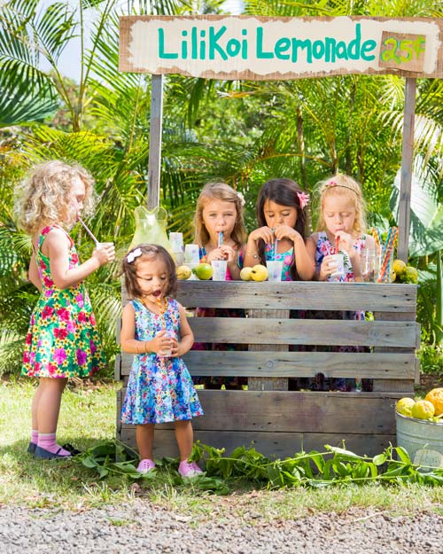 5 girls wearing Hawaiian print Holoholo Dresses all drink lemonade from straws at a rustic Maui style Lilikoi Lemonade stand.