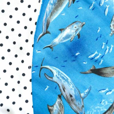 Close up view of Baby Blue by Cool Blue Maui’s Swim with Dolphins Romper which has an ocean blue background with grey swimming dolphins and reverses to a white with black polkadot fabric.